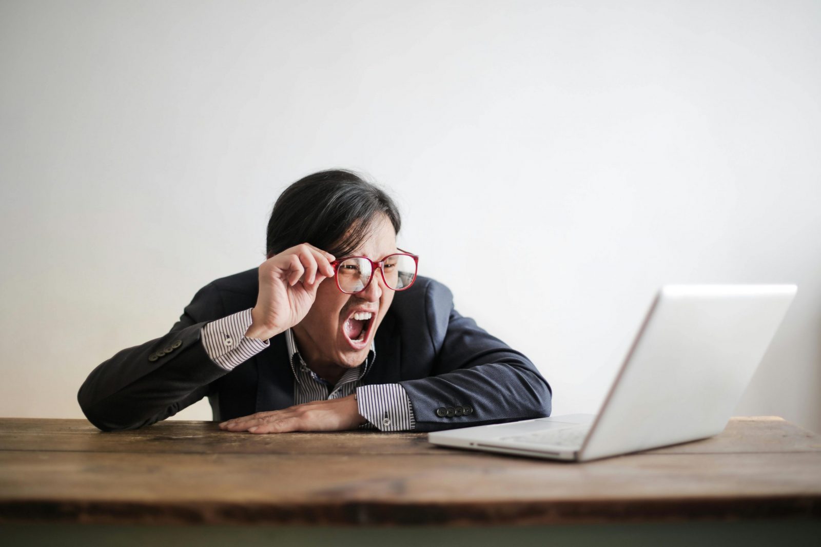 Modern Asian man in jacket and glasses looking at laptop and screaming with mouth wide opened on white background